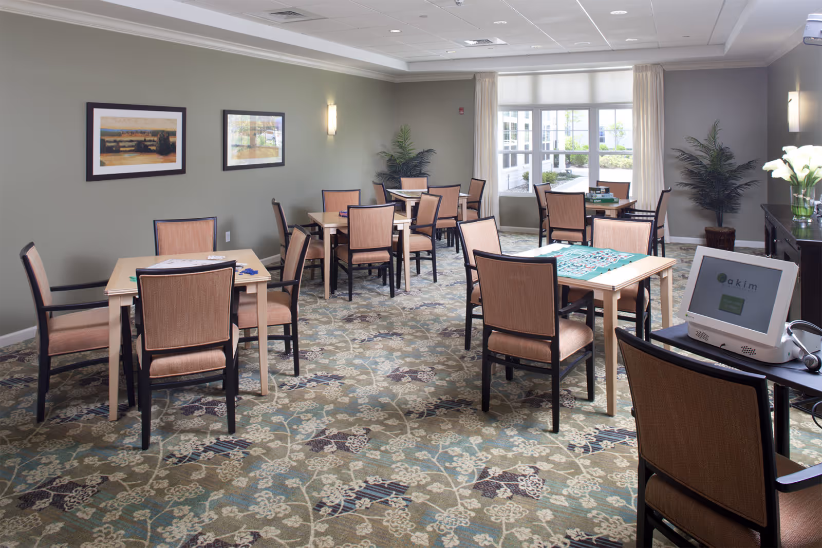 A well-lit activity room with several tables and chairs arranged for group activities. The tables have board games and game pieces on them. The room has a large window with curtains, framed artwork on the walls, potted plants, and a patterned carpet. A computer monitor is visible on a table in the foreground.