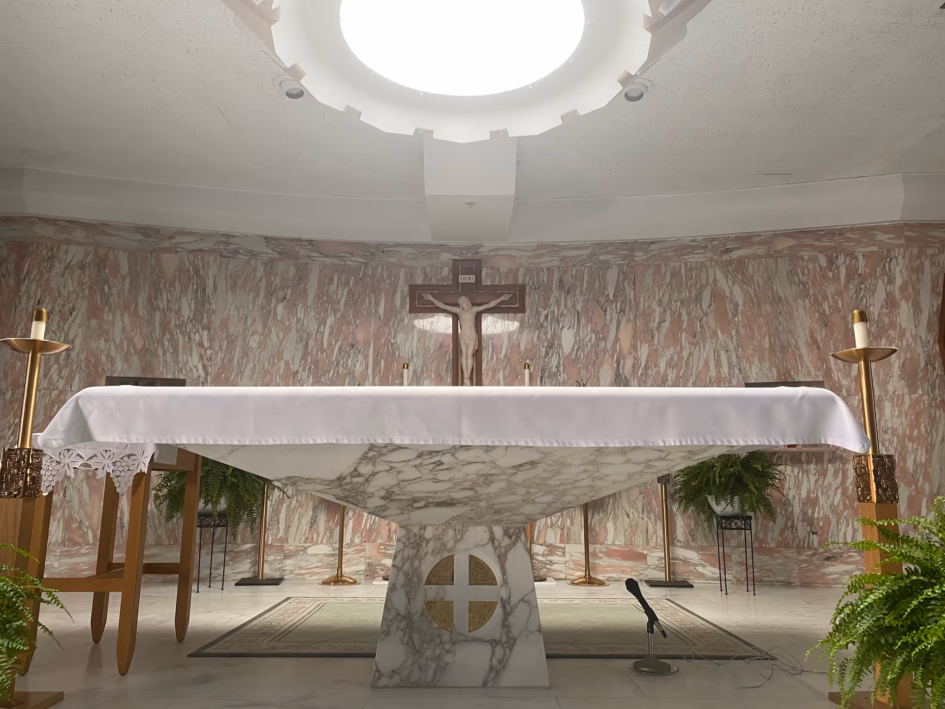 Interior view of a chapel with a marble altar covered by a white cloth, a crucifix mounted on the marble wall behind the altar, two tall gold candle holders on either side, and green plants placed around the altar area.