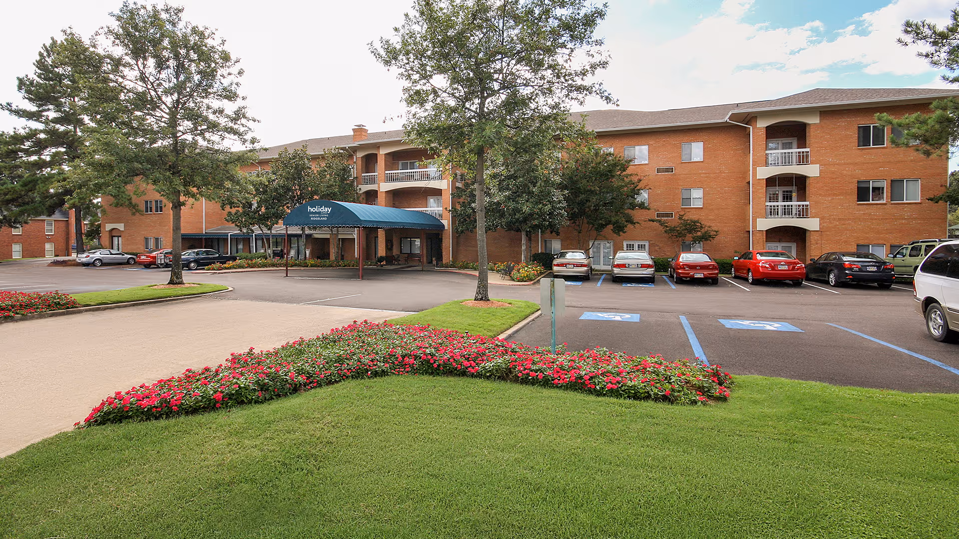 Exterior view of Holiday Chateau Ridgeland, a three-story brick building with balconies and a blue awning over the entrance. The foreground features a well-maintained lawn with flower beds and trees, and a parking lot with several cars, including handicap parking spaces.