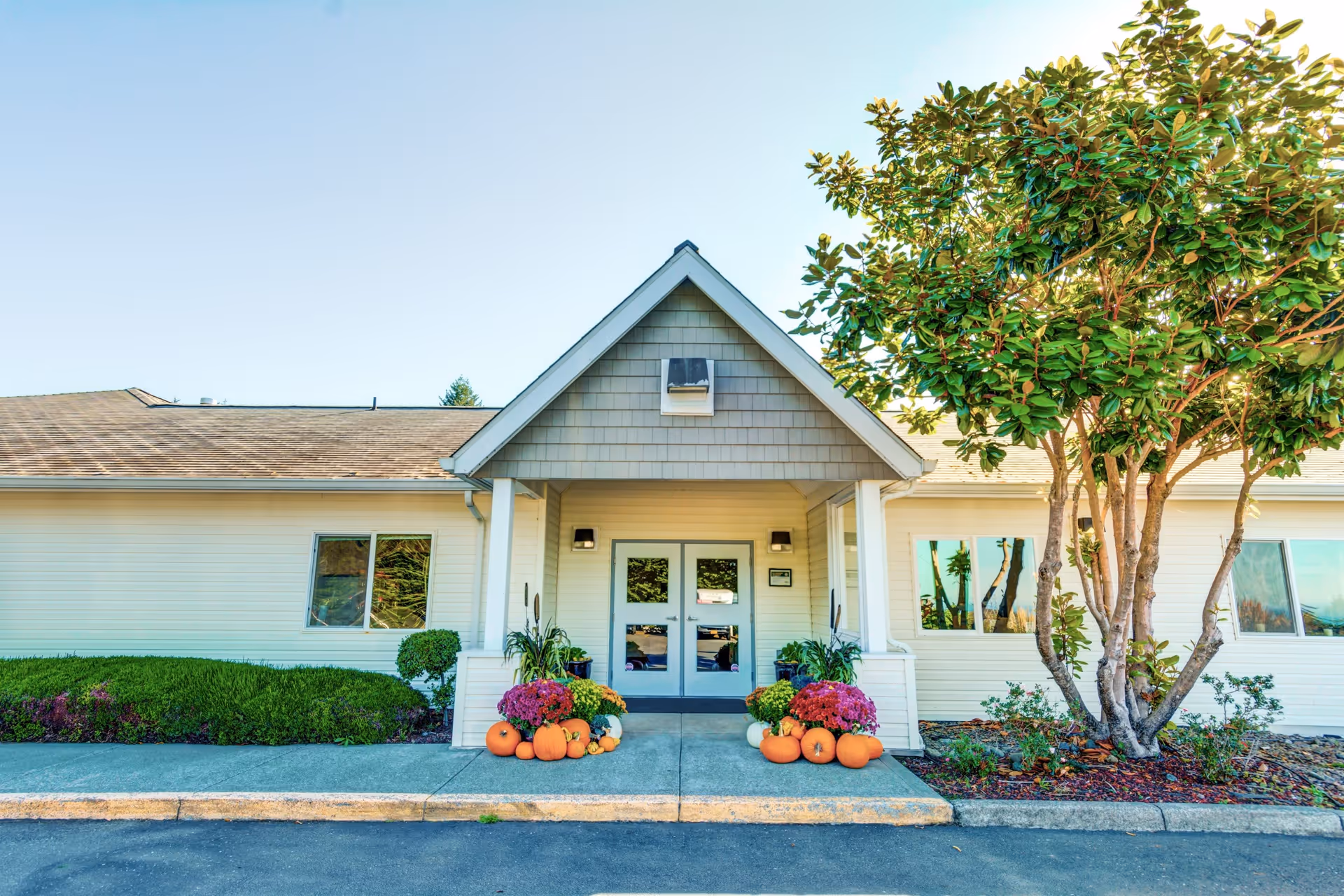 Front entrance of a single-story building with a peaked roof over the doorway. The entrance is decorated with orange pumpkins and colorful flowers on both sides. There are shrubs and a tree to the right of the entrance, and the sky is clear and blue.