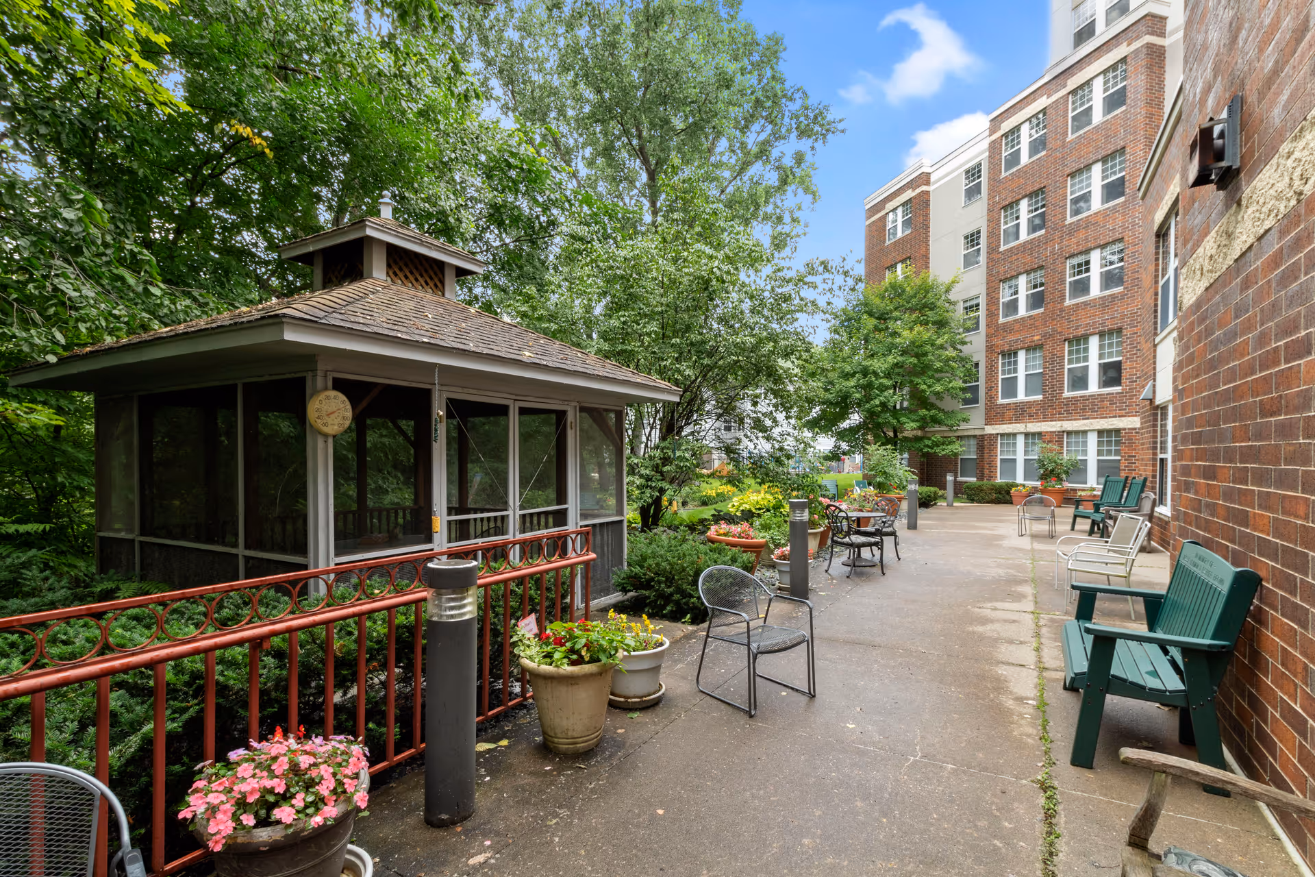 Outdoor patio area at Eaglecrest Senior Housing featuring a screened gazebo, various chairs and benches, potted plants, and a brick building with multiple windows in the background under a partly cloudy sky.