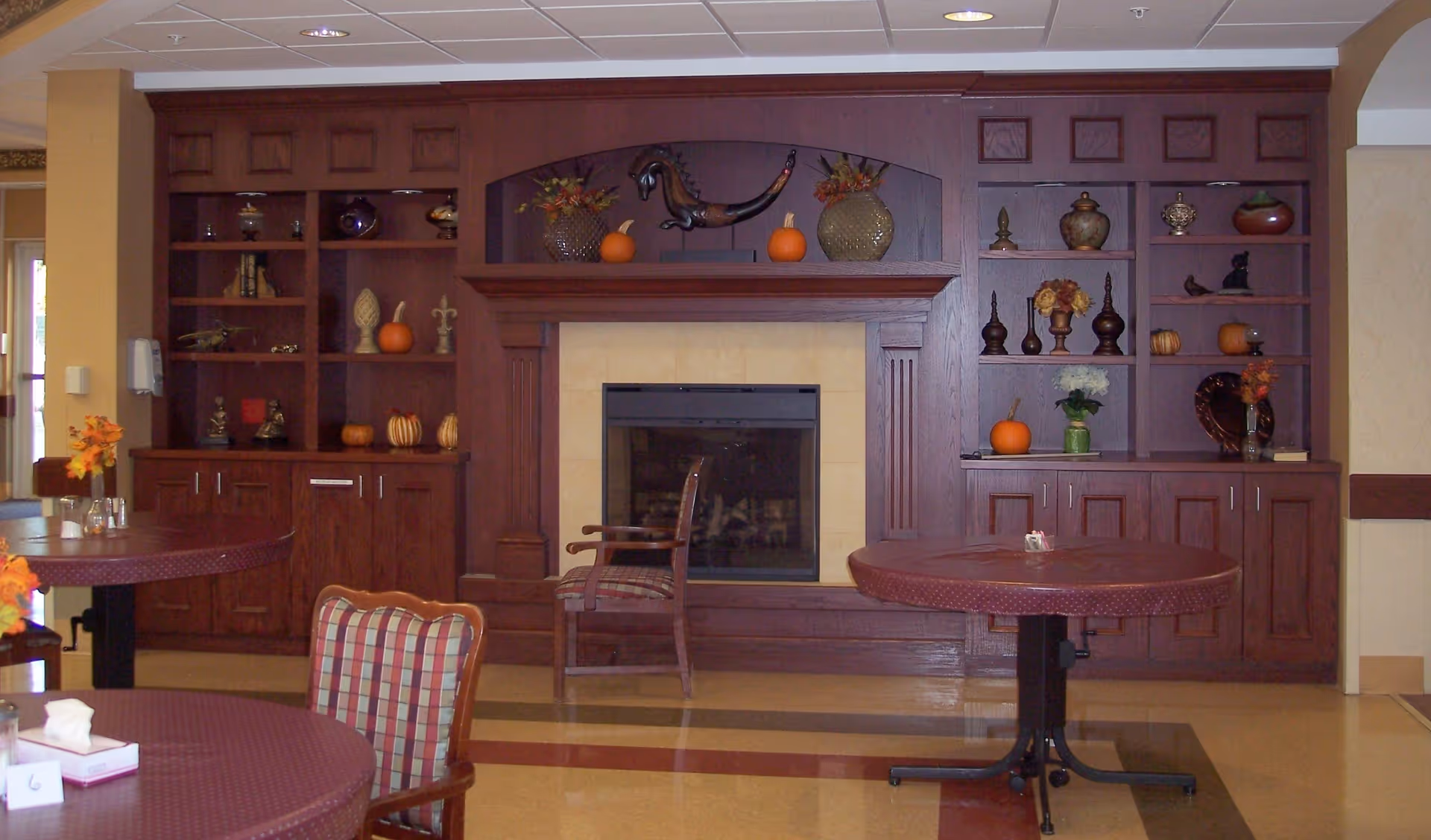 A cozy interior room featuring a wooden fireplace with built-in shelves on either side, decorated with small pumpkins, vases, and ornamental items. In front of the fireplace, there is a wooden chair. The room also contains round tables with patterned tablecloths and cushioned chairs, suggesting a common or lounge area.
