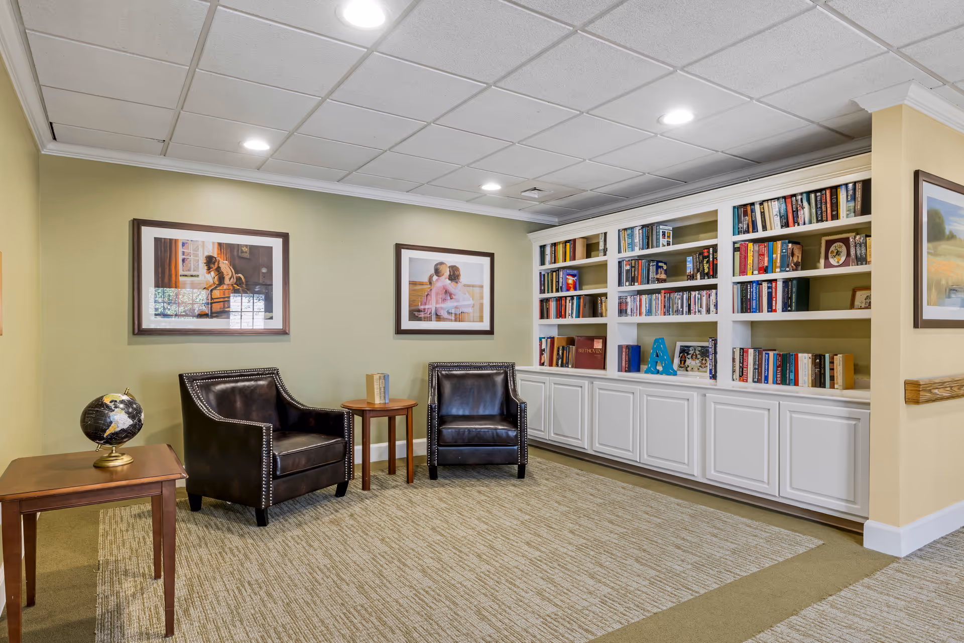 A cozy reading nook in a senior living facility featuring two dark leather armchairs with nailhead trim, a small round wooden side table between them, and a wooden table with a globe on it. Behind the chairs is a built-in white bookshelf filled with books and decorative items. The walls are painted light green and adorned with framed artwork. The floor is carpeted with a patterned rug, and the ceiling has recessed lighting.