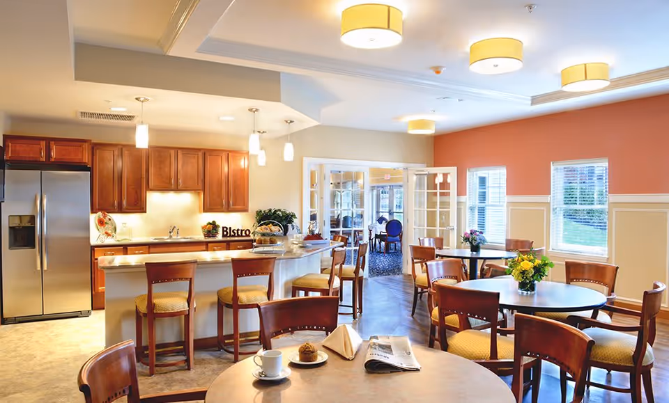 Bright communal dining area with a kitchen island and bar stools, round dining tables and chairs, and pendant and ceiling lights.