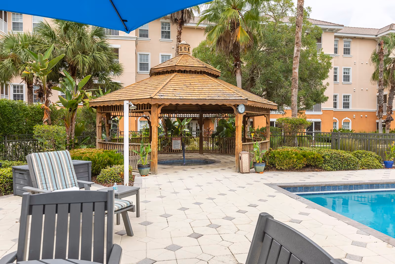Outdoor pool area with a wooden gazebo surrounded by greenery and palm trees. There are lounge chairs with striped cushions and a blue umbrella providing shade. A multi-story building is visible in the background.