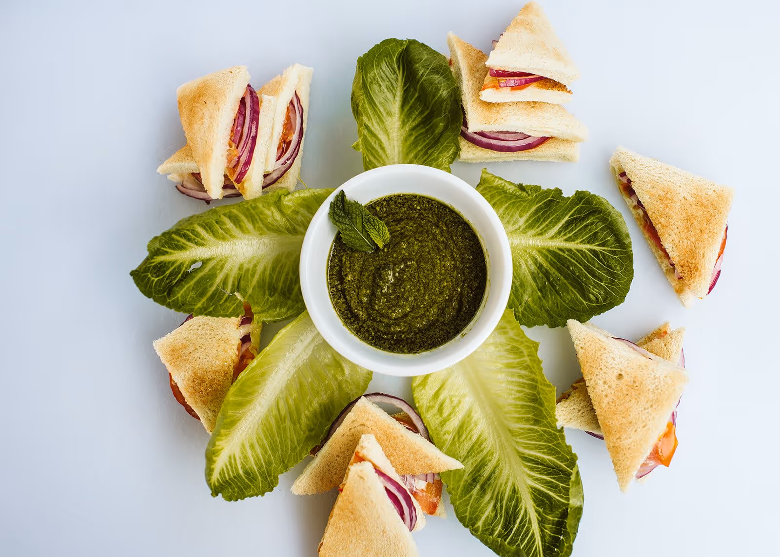 A white bowl filled with green pesto sauce garnished with a mint leaf, surrounded by fresh romaine lettuce leaves and small triangular sandwiches with toasted bread, tomato, and red onion on a white surface.