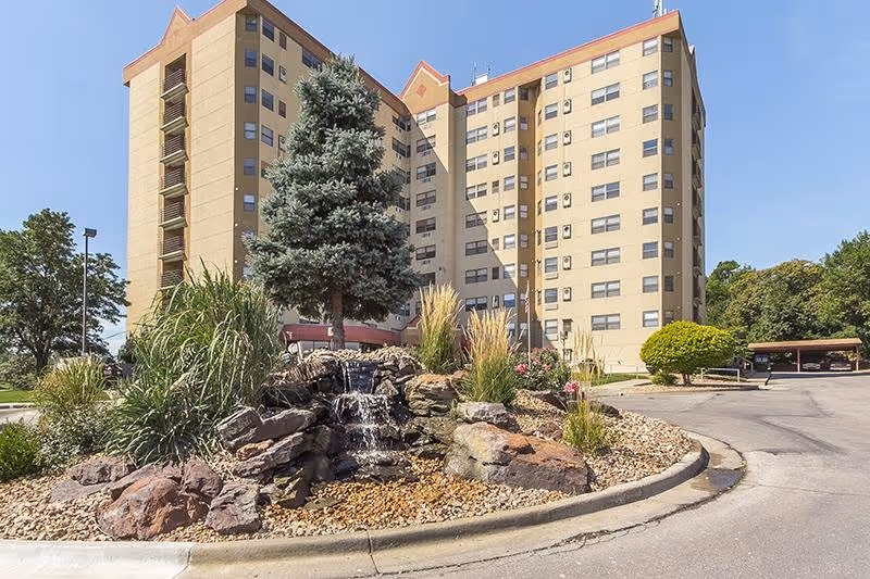 Exterior view of a multi-story senior living community building with a landscaped roundabout featuring rocks, a small waterfall, a pine tree, and various plants under a clear blue sky.