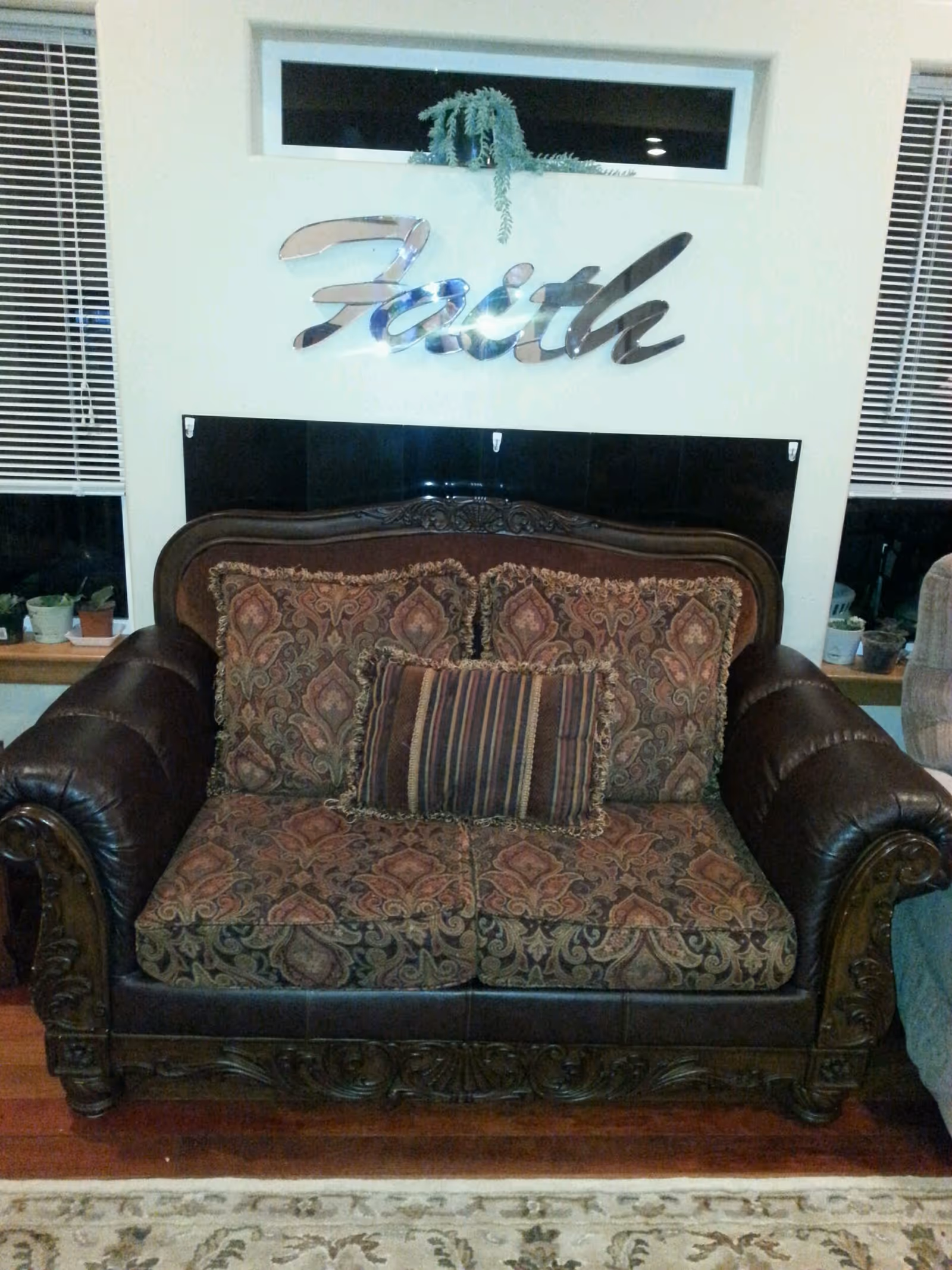 A patterned loveseat with dark leather armrests and carved wooden details sits against a wall. Above the loveseat is a shiny metallic wall decoration spelling the word 'Faith'. There are two windows with blinds on either side of the loveseat, and small potted plants are placed on the window sills.