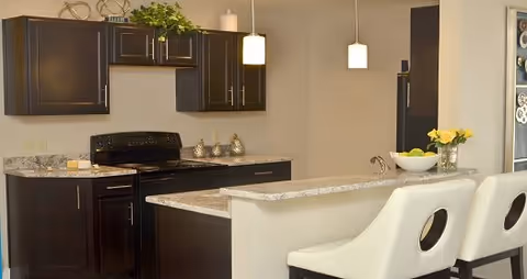 Modern kitchen with dark wood cabinets, granite countertops, a black stove, and a kitchen island with two white bar stools. Pendant lights hang above the island, and there are decorative items including a bowl of lemons and a vase with yellow flowers.