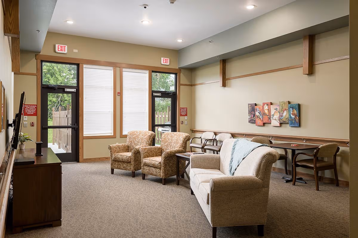 A senior living facility common area with beige walls and carpeted floor. The room features a beige sofa with a light blue throw blanket, two patterned armchairs, a wooden side table, and a TV mounted on the wall opposite the seating. There are two exit doors with windows and blinds, and a small table with chairs along the right wall beneath colorful bird artwork.
