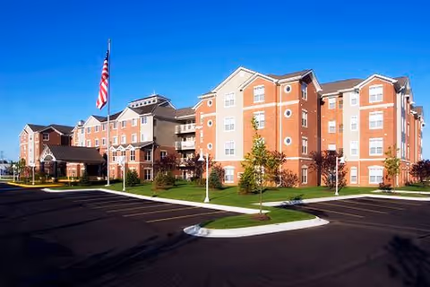 Exterior view of Northpoint Village of Utica, a brick senior living building with flag and parking.