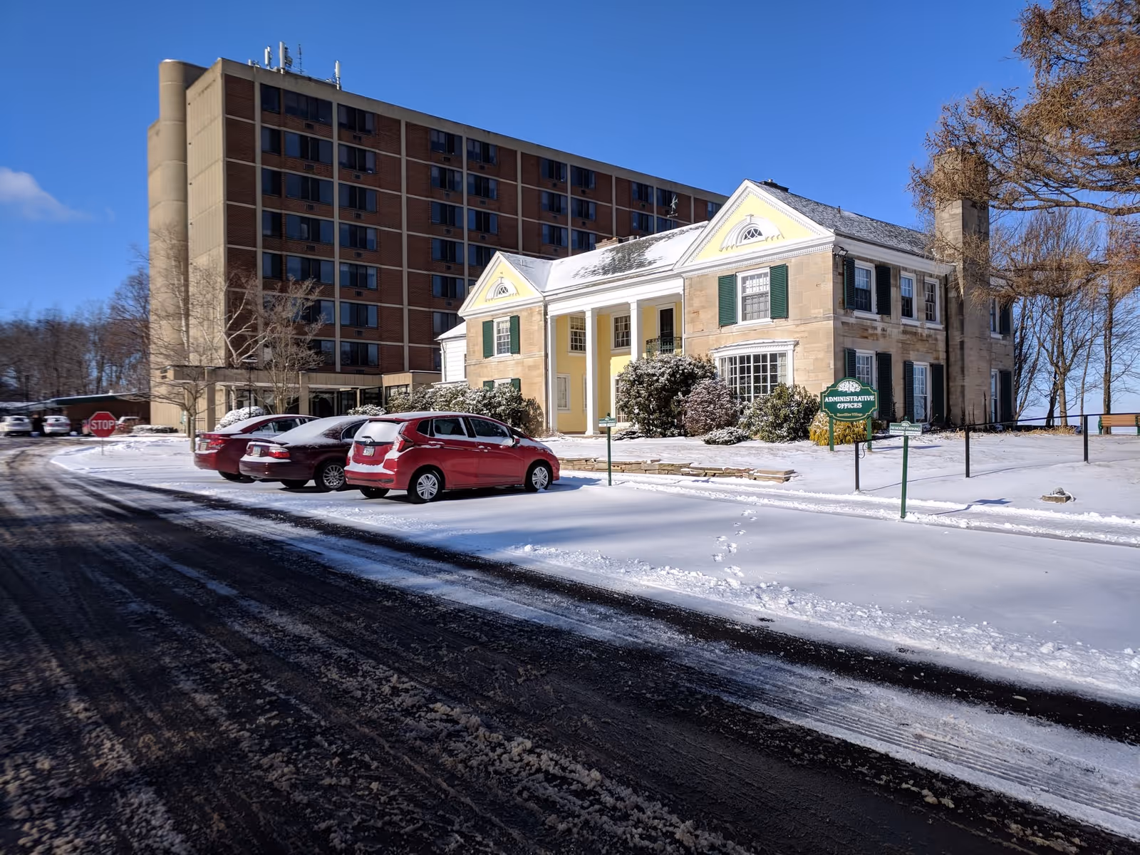Snow-covered parking lot with several cars parked next to a large multi-story brick building and a smaller historic-style building labeled as Administrative Offices at Brevillier Village, under a clear blue sky.