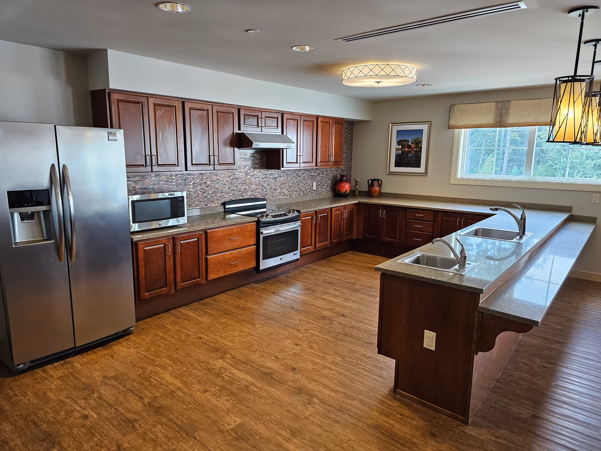 A spacious kitchen with wooden cabinets, a stainless steel refrigerator, microwave, stove with range hood, and a large countertop island with two sinks. The kitchen has wood flooring, a window with a beige blind, and decorative vases on the countertop.