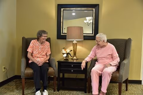 Two elderly women sitting in high-backed armchairs facing each other in a warmly lit room. Between them is a small black side table with a gold lamp and a small flower arrangement. A large mirror hangs on the wall behind the table, reflecting part of a chandelier.