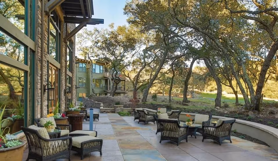 Outdoor patio area at Fountaingrove Lodge with multiple wicker chairs and small tables arranged on a tiled floor. The patio is adjacent to a building with large windows and surrounded by trees and greenery under a clear sky.