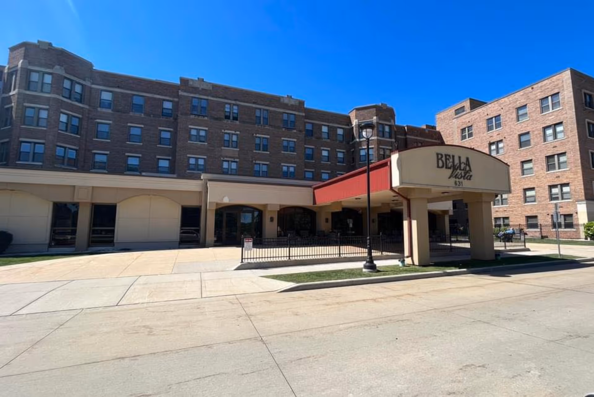 Exterior view of a multi-story brick building under a clear blue sky. The building has multiple windows and a covered entrance with a red awning. A sign on the awning reads 'Bella Vista 631'. There is a sidewalk and street in the foreground.