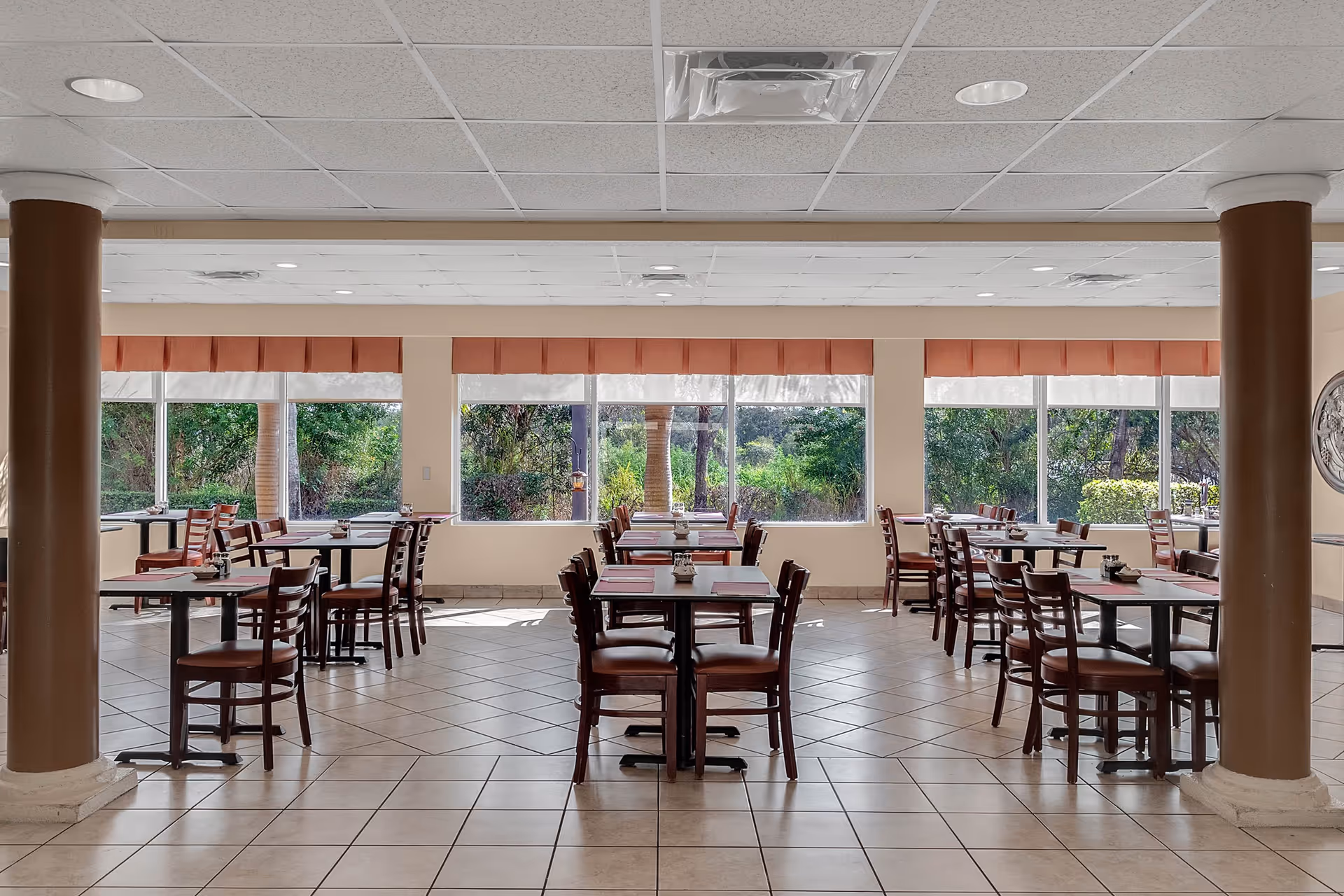 A bright dining room with multiple wooden tables and chairs arranged neatly on a tiled floor. Large windows along the back wall let in natural light and offer a view of green trees and shrubs outside. The ceiling has recessed lighting and two large brown columns are visible in the foreground.