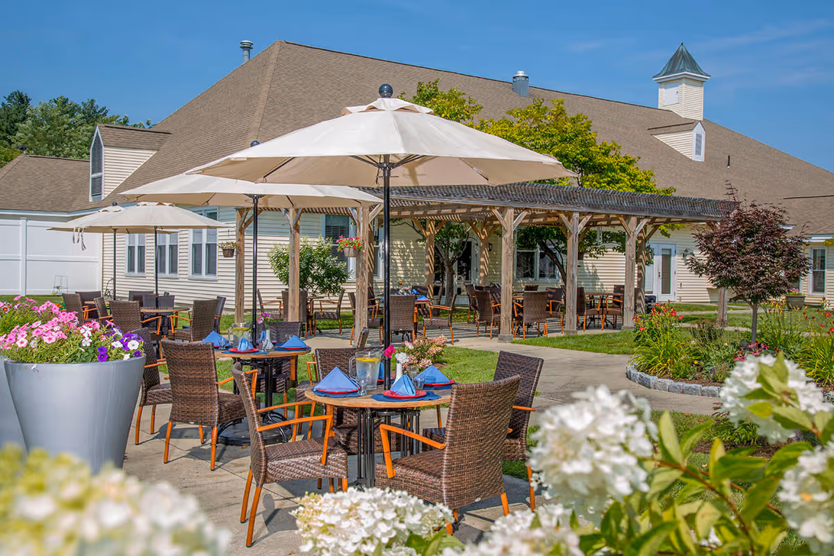 Outdoor patio area at The Arbors at Shelburne with tables and chairs arranged under large white umbrellas. The setting includes a wooden pergola, colorful flowers in planters, and a building with beige siding in the background under a clear blue sky.