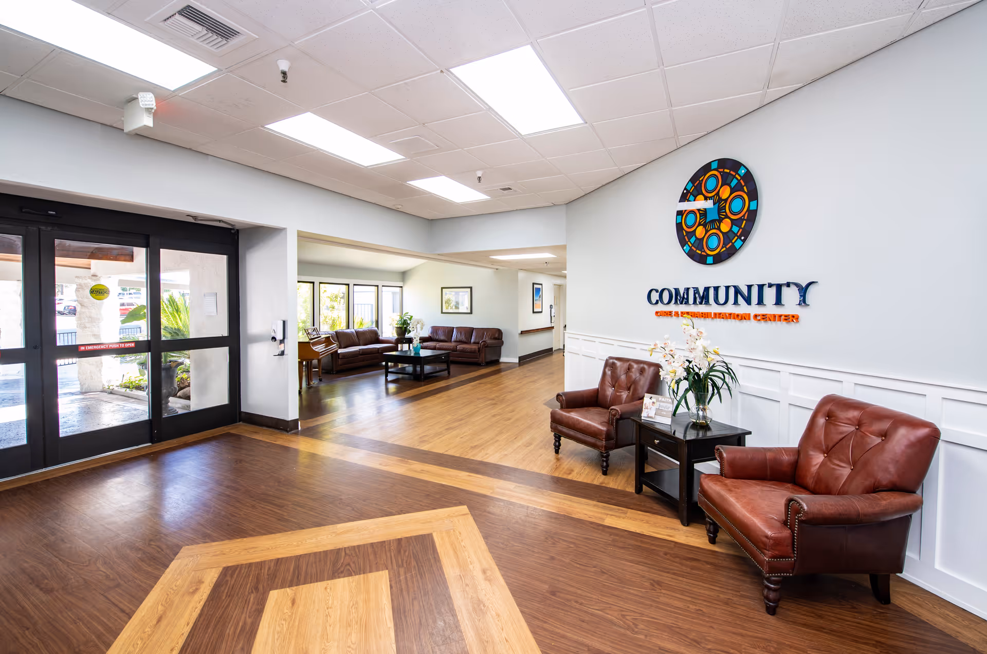 Interior view of the Community Care & Rehabilitation Center lobby area featuring a seating area with two brown leather chairs and a small table with flowers. The floor has a wood pattern, and there are large glass doors to the left. In the background, there is another seating area with sofas and a coffee table near windows.