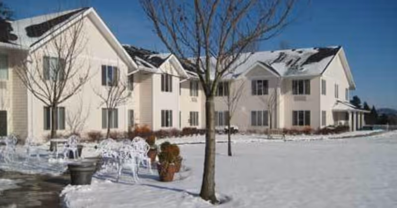 Exterior view of a two-story assisted living facility building named Somerset Assisted Living, surrounded by snow-covered ground, leafless trees, and outdoor white metal chairs and tables.