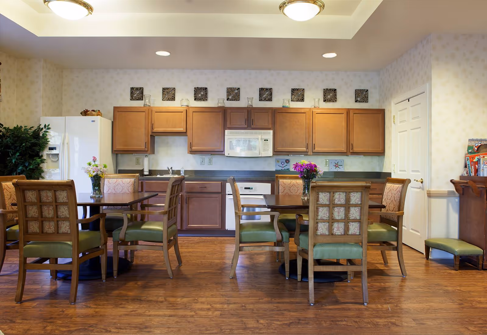 A cozy dining area with two wooden tables, each surrounded by four cushioned chairs with green seats and patterned backs. The room features a kitchen area in the background with wooden cabinets, a white refrigerator, microwave, and dishwasher. There are decorative items on top of the cabinets and fresh flowers in vases on each table. The floor is wooden, and the walls have a light patterned wallpaper.