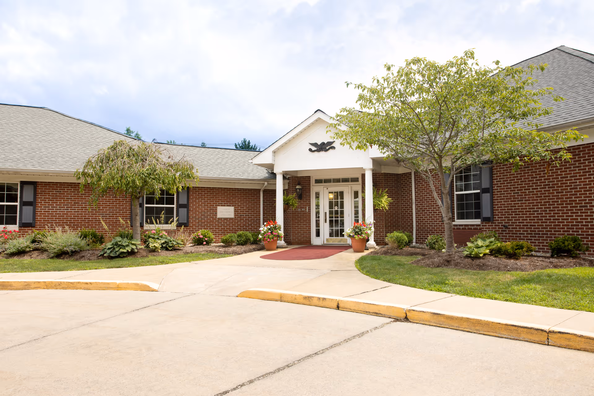 Front exterior view of a single-story brick building with a gray shingled roof, white columns at the entrance, and a red carpet leading to double glass doors. There are two trees and various plants and flowers in landscaped beds near the entrance under a partly cloudy sky.