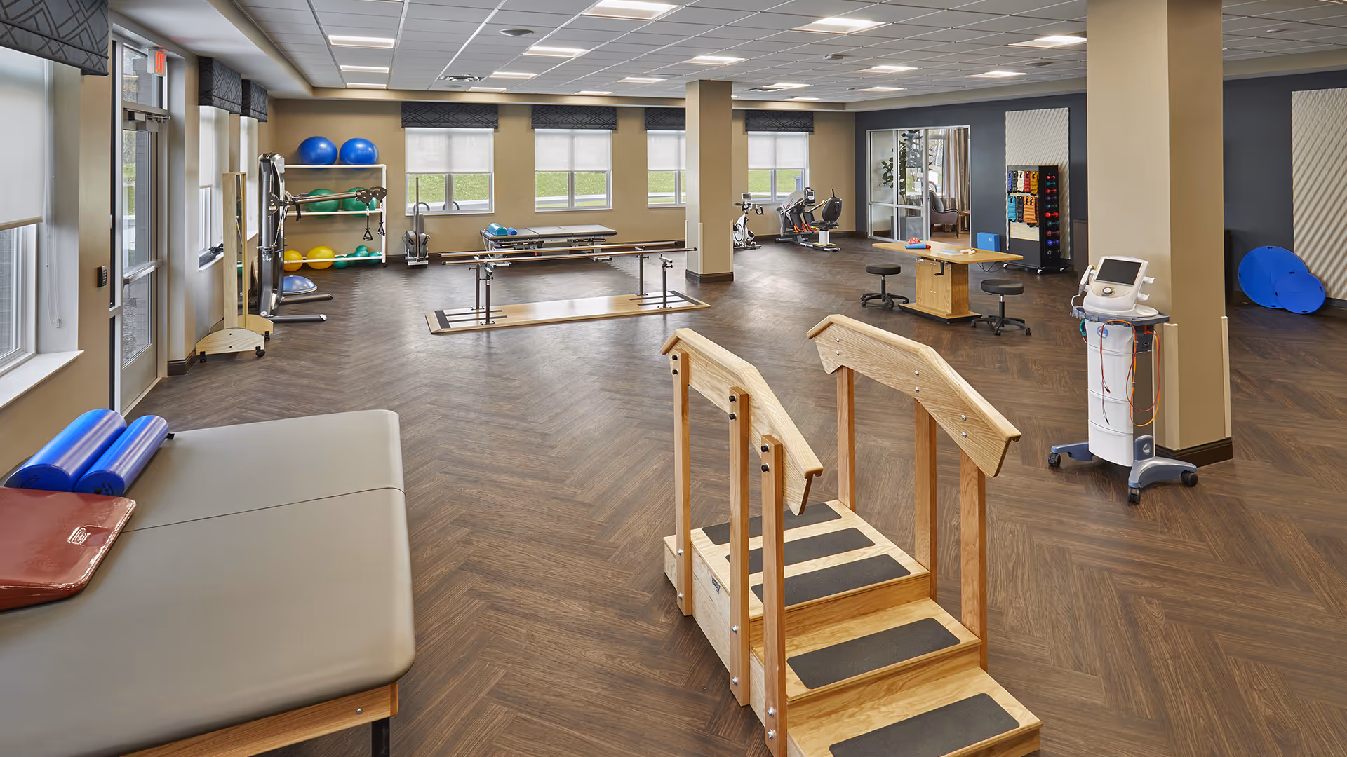 A spacious physical therapy or rehabilitation room with wooden flooring and beige walls. The room contains various exercise and therapy equipment including parallel bars, therapy steps, exercise balls, a treatment table, stationary bikes, and a therapy machine. Large windows with blinds allow natural light to fill the room.
