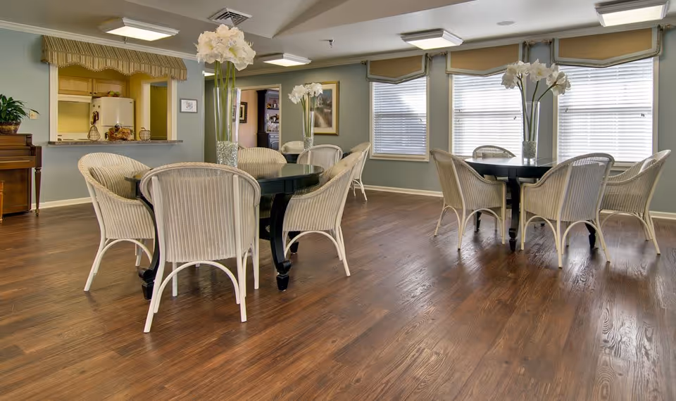 A bright and spacious dining area with two round black tables, each surrounded by six white wicker chairs. Each table has a tall glass vase with white flowers. The room has wooden flooring, large windows with blinds, and a pass-through window to a kitchen area. The walls are painted light blue with framed artwork and a piano is visible on the left side.