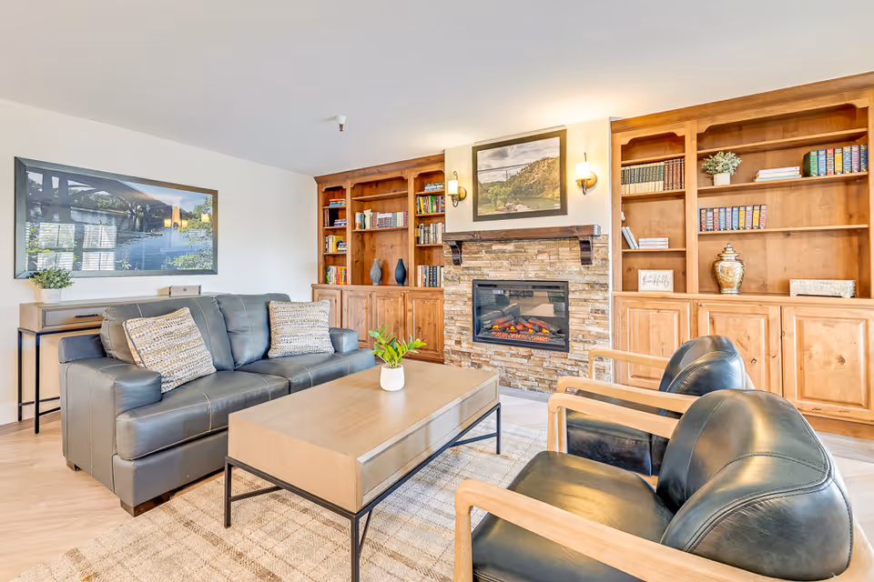 A cozy living room with a gray leather sofa, two black leather armchairs with wooden frames, a light wood coffee table with a small plant, built-in wooden bookshelves filled with books and decorative items, a stone fireplace with a framed picture above it, and a large framed photo on the wall.