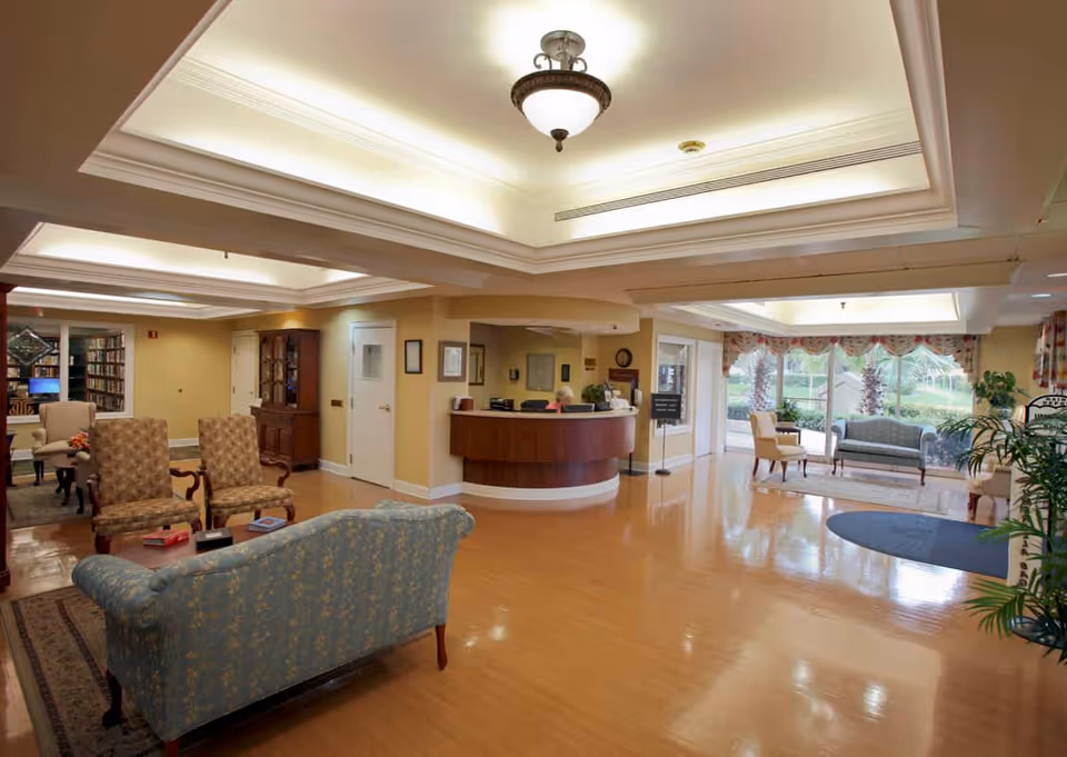 A spacious and well-lit common area in a senior living facility with comfortable seating including armchairs and sofas arranged around small tables. There is a reception desk with a staff member behind it, large windows with floral curtains letting in natural light, and a bookshelf visible in the background. The floor is polished wood, and the ceiling has recessed lighting and a central light fixture.