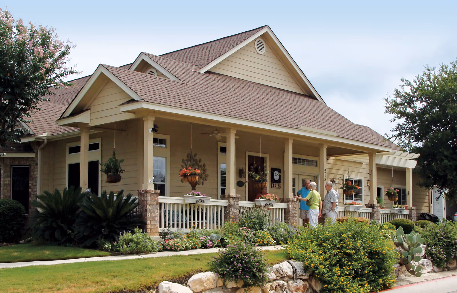 Single-story beige building with a covered front porch, landscaped yard, and three people talking on the porch.