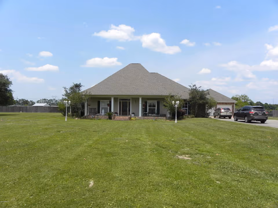 Single-story ranch-style house with a wide front porch, large grassy lawn, and cars parked at the driveway under a blue sky.