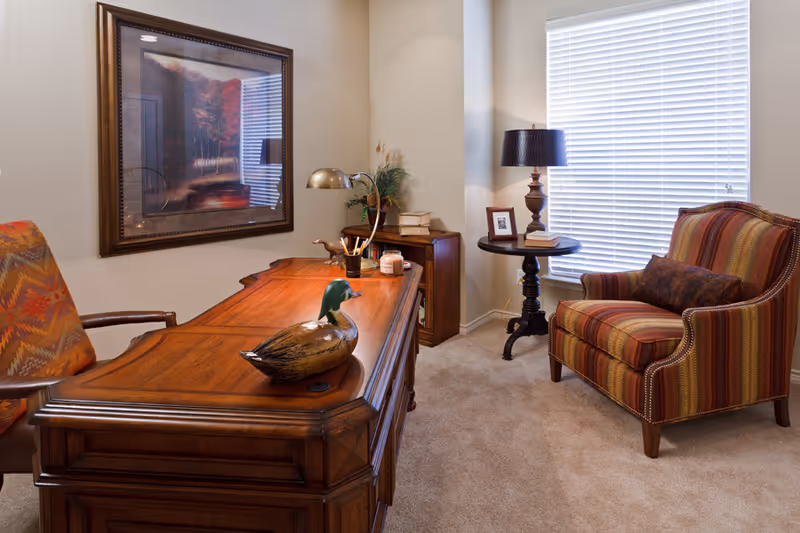 A cozy interior room with a wooden desk featuring a decorative duck figurine, a desk lamp, and some stationery. There is a patterned armchair on the left and a striped upholstered armchair with a dark cushion on the right near a window with white blinds. A small round side table with a lamp, a framed photo, and a book is positioned between the armchair and the window. A framed painting hangs on the wall above the desk.