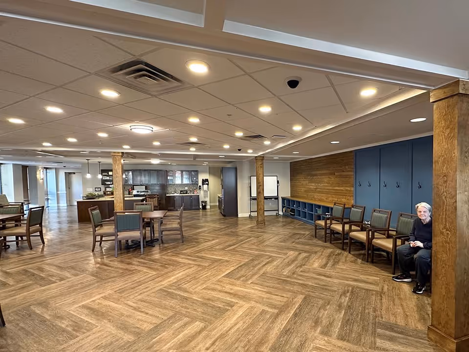 Spacious senior living common area with herringbone wood floors, tables and chairs, a kitchenette in the background, and a person seated along the right wall.