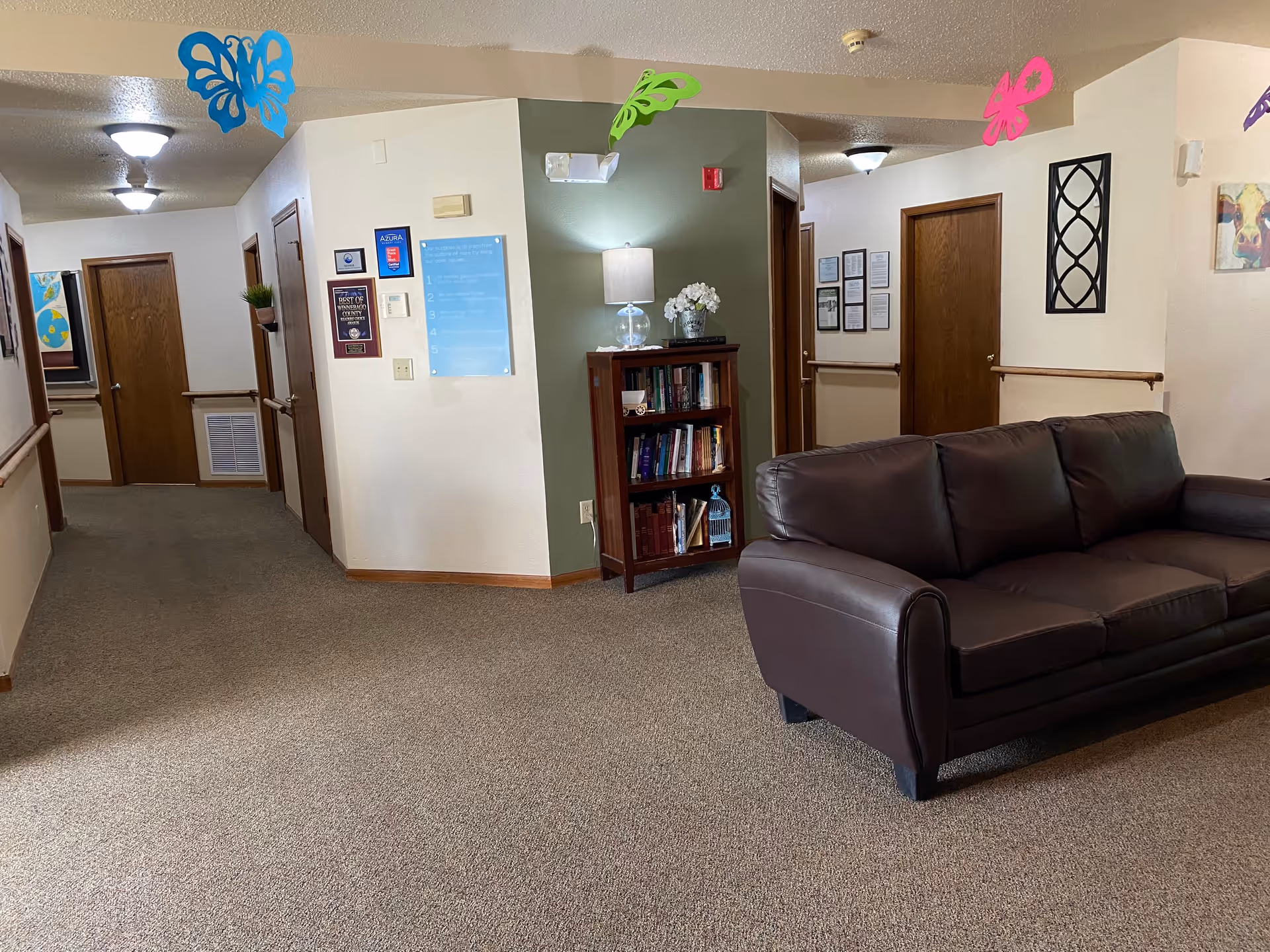Interior hallway of a senior living facility with a brown leather couch on the right, a wooden bookshelf with a lamp and flowers, several closed wooden doors, framed pictures and plaques on the walls, and colorful butterfly decorations hanging from the ceiling.