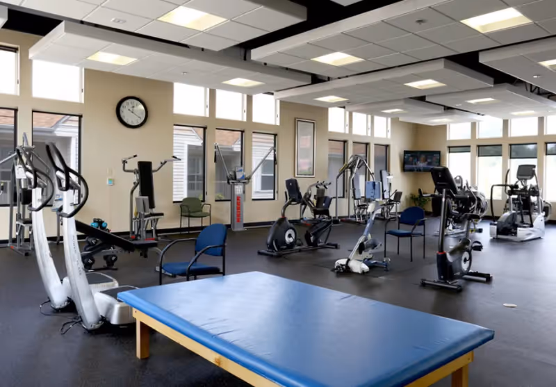 A spacious fitness room with various exercise equipment including stationary bikes, elliptical machines, weight machines, and a large blue padded exercise table in the foreground. The room has large windows allowing natural light, beige walls, and a wall clock.