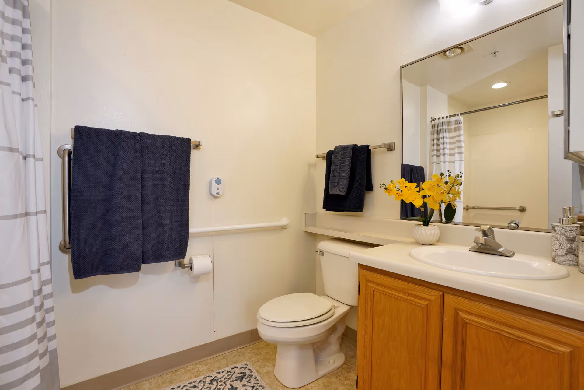 A clean bathroom with a toilet, a sink with a wooden cabinet, a large mirror, and a shower with a striped curtain. Two dark blue towels hang on metal towel racks, and a small vase with yellow flowers is placed on the countertop.
