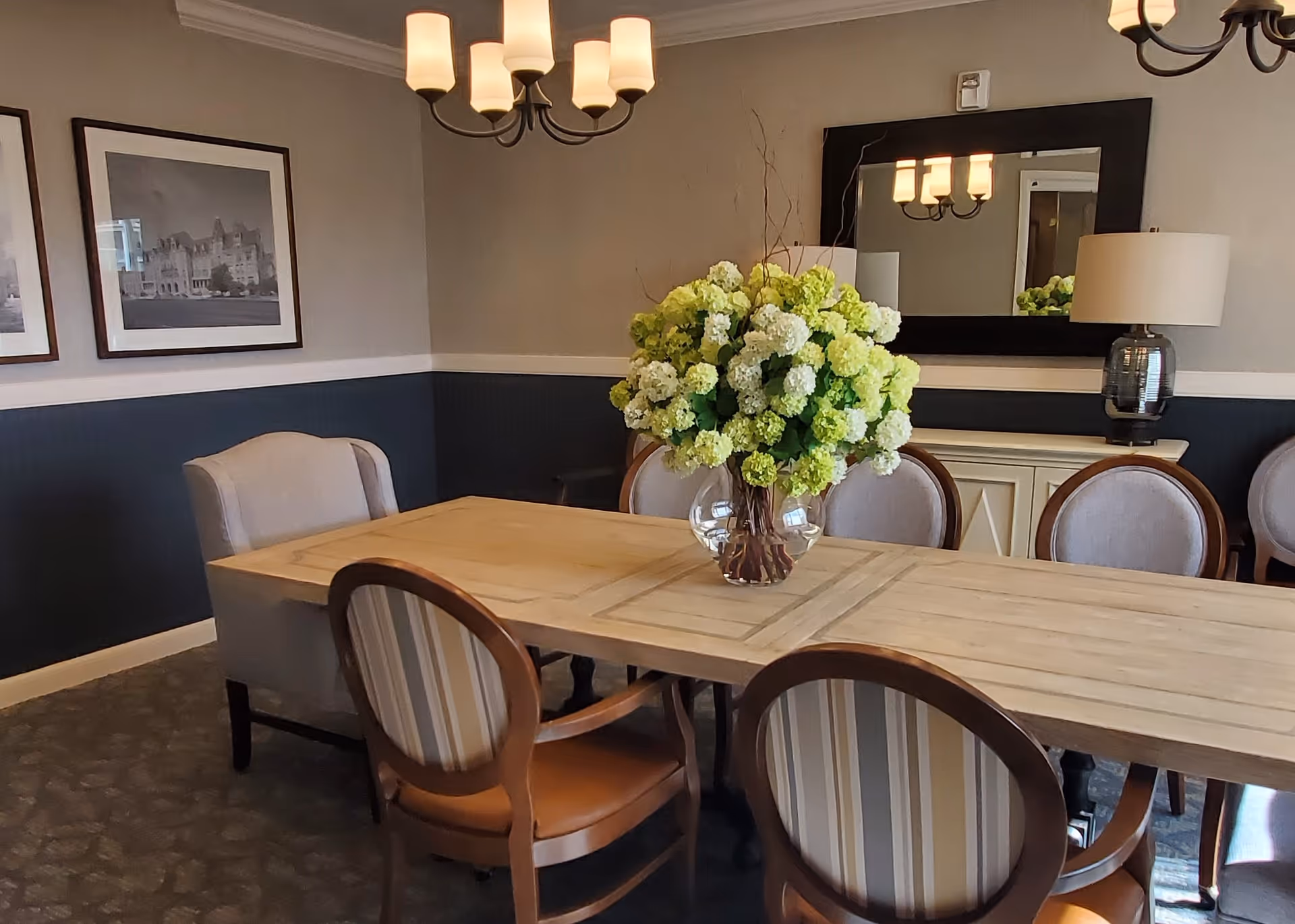 A dining room with a wooden table surrounded by six chairs, a large vase with white and green flowers in the center of the table, a large mirror on the wall, a table lamp, and two framed black and white photographs on the wall.