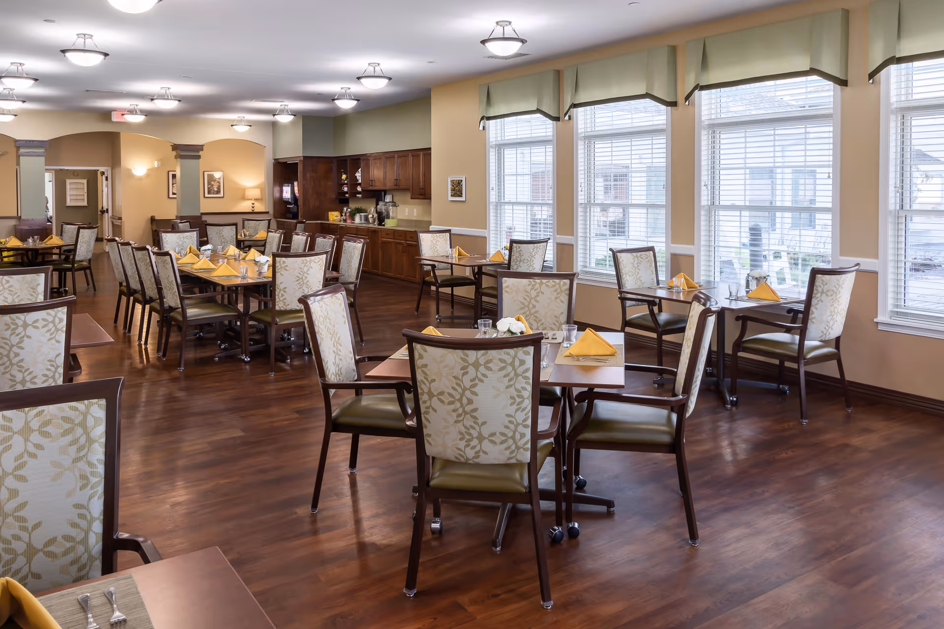 A bright and spacious dining room with multiple tables set with yellow folded napkins, glasses, and silverware. The room features large windows with blinds, wooden flooring, and comfortable chairs with patterned upholstery. There is a counter area with cabinets and a coffee machine in the background.