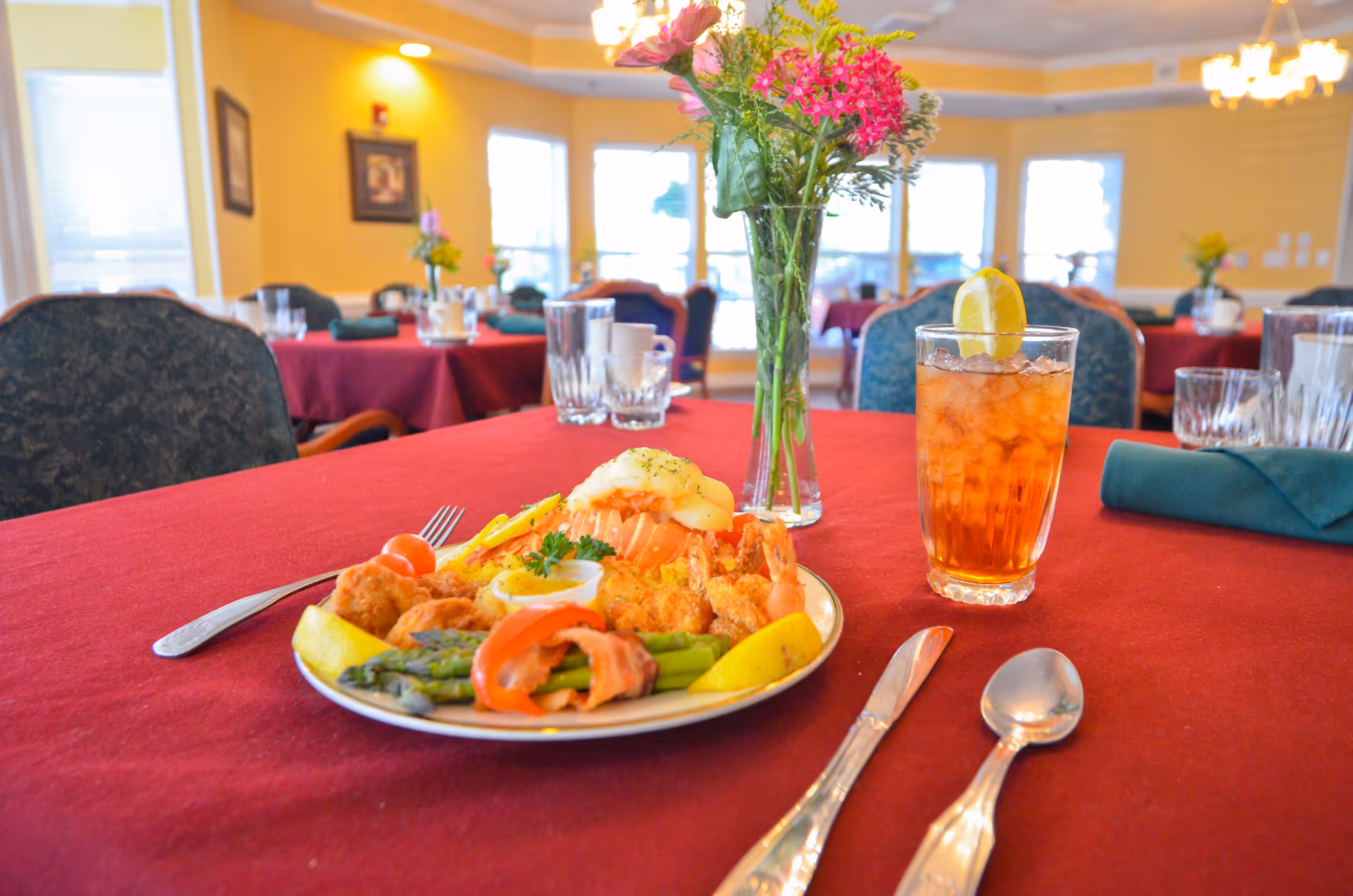 A dining table with a red tablecloth set with a plate of food including asparagus wrapped in bacon, fried shrimp, a lemon wedge, and a glass of iced tea with a lemon slice. The background shows a dining room with several tables covered in red tablecloths, chairs, and windows letting in natural light. A vase with pink and yellow flowers is also on the table.