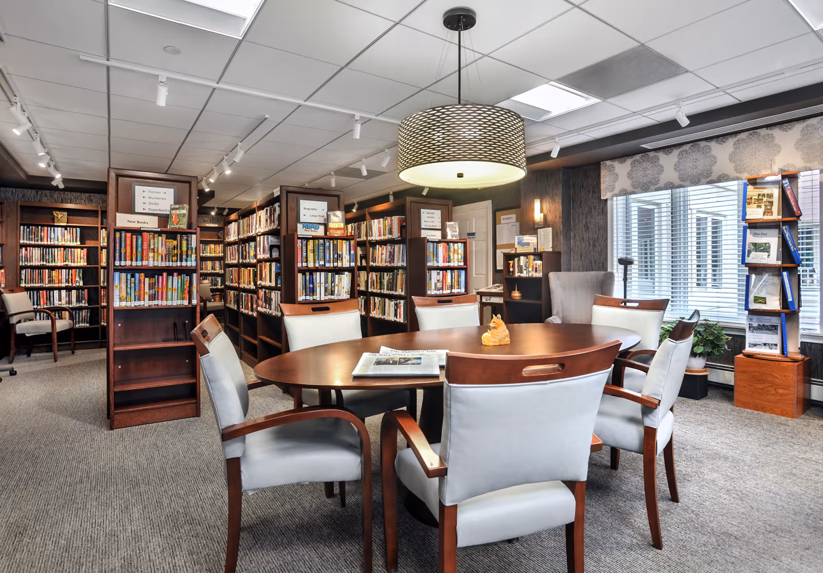 A cozy library room with multiple bookshelves filled with books, a round wooden table surrounded by six cushioned chairs, a large circular hanging light fixture above the table, and a window with blinds and patterned valance. There are also two wooden display stands with brochures near the window and a comfortable armchair in the corner.