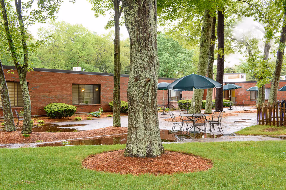 Outdoor courtyard with trees, wet pavement, patio tables and umbrellas in front of a single-story brick building.