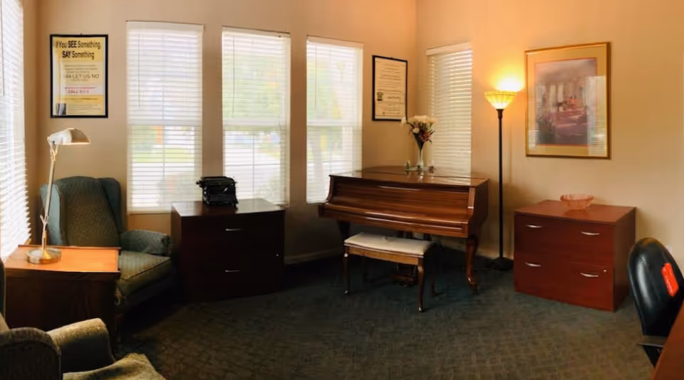 A cozy room with three large windows covered by white blinds, a wooden piano with a cushioned bench, two wooden filing cabinets, a green upholstered armchair, a floor lamp, a table lamp, and framed pictures on the walls.