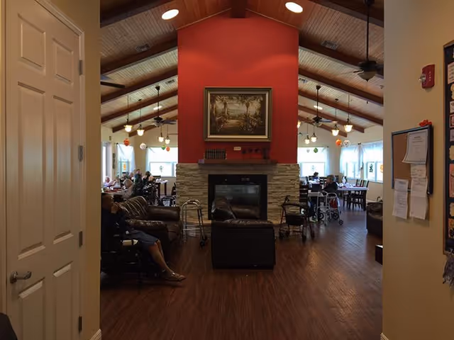 Open common room in an assisted living facility with a central fireplace, seating area, and dining tables under a vaulted ceiling.