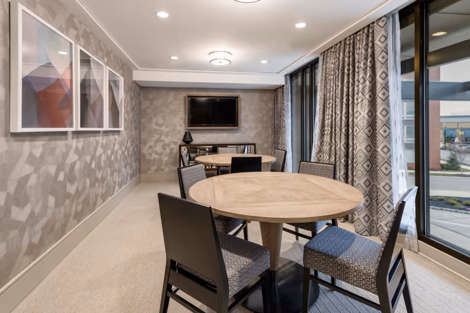 A modern dining area with two round wooden tables surrounded by upholstered chairs. The room features patterned wallpaper, three abstract framed artworks on one wall, a flat-screen TV mounted on the far wall, and large windows with patterned curtains letting in natural light.