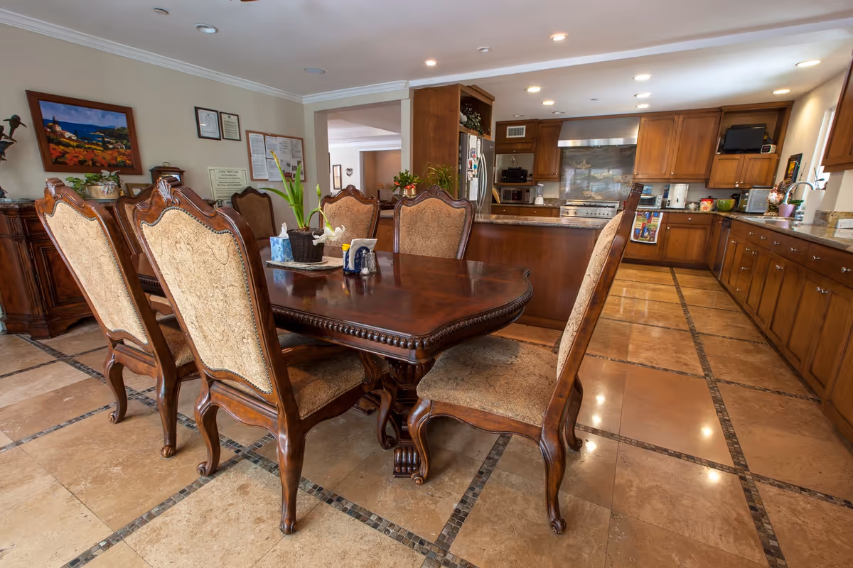 Ornate wooden dining table with upholstered chairs in a spacious room opening to a large kitchen with wooden cabinets and tiled floor.