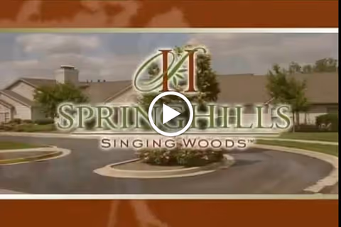 Entrance view of the Spring Hills Singing Woods senior living facility with a circular driveway and landscaped greenery in front of a single-story building under a partly cloudy sky.