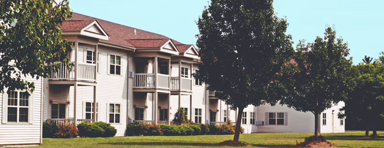 Exterior view of a two-story residential building with white siding and red roof, featuring balconies and surrounded by green trees and grass under a clear sky.