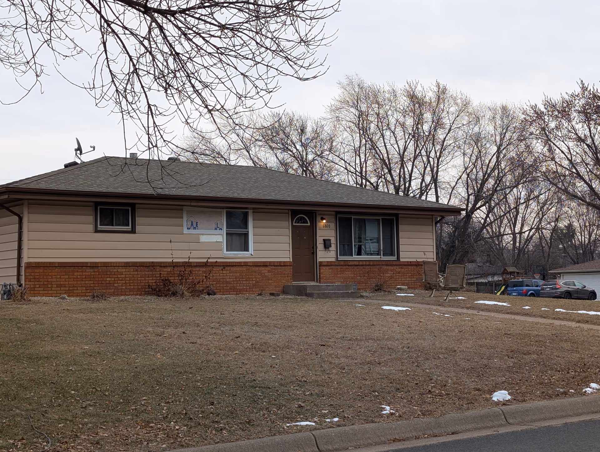 Single-story house with beige siding and brick lower exterior, a brown front door, and two windows. The house is set on a lawn with patches of snow and leafless trees in the background under an overcast sky.