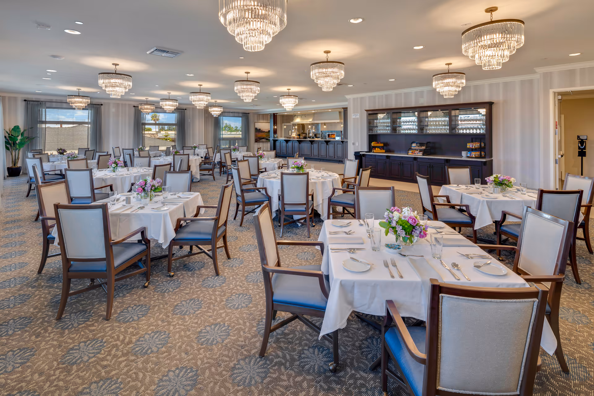 A spacious and elegant dining room with multiple round and square tables covered with white tablecloths, each set with plates, glasses, silverware, and floral centerpieces. The room features patterned carpet, large windows with sheer curtains, and several ornate chandeliers hanging from the ceiling. A dark wooden buffet cabinet with glassware and snacks is visible along one wall, and a serving counter is seen in the background.